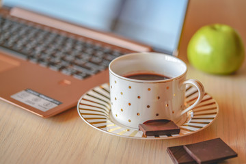 Tempting cup of coffee on the wooden table with laptop and apple on background. Piece of dark chocolate is on the saucer, break time in the office for refreshment, closeup