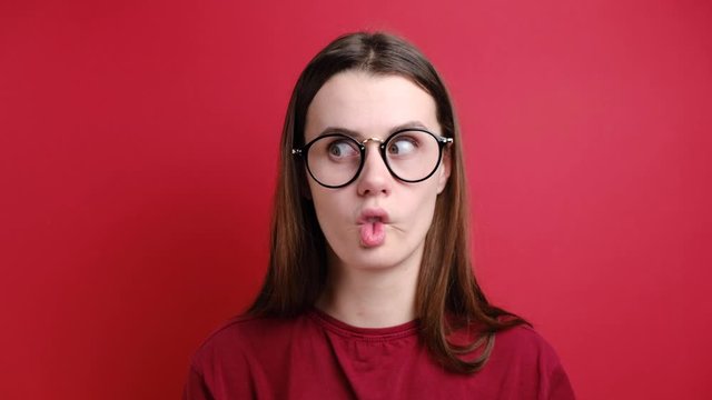 Crazy comic female in glasses makes funny face, crosses eyes, plays fool, doesnt want to be responsible, wears red t-shirt and stands over red background. Facial expressions. 