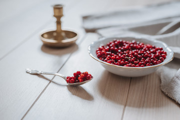 Close up of lingonberries in a bowl and on a spoon, linen tablecloth and candlestick on wooden table in bright sunlight, rustic style with grain