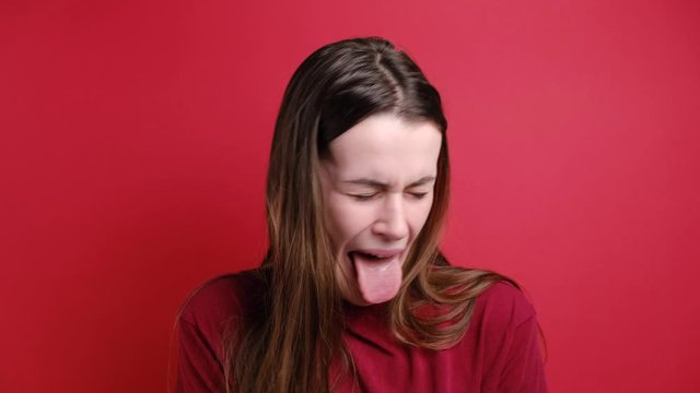 Unhappy woman frowns face, has disgusting expression, shows tongue, irritated with somebody, dressed in  t-shirt, isolated over red background. People and negative facial expressions