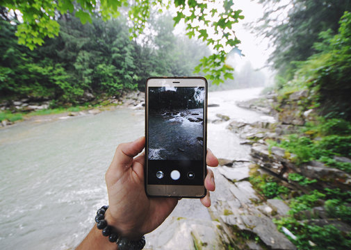 Young man hipster makes photo of mountain and river landscape on smartphone camera, to share on internet social media through photo application for mobile device.