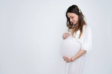 Beautiful brunette pregnant woman with loose hair dressed in white underwear stands with closed eyes and holds her hands on her swollen belly