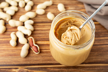 Creamy peanut paste in open glass jar, peanut butter in spoon. Peanuts in the peel scattered on the brown wooden table with copy space for cooking breakfast. Vegan food concept.