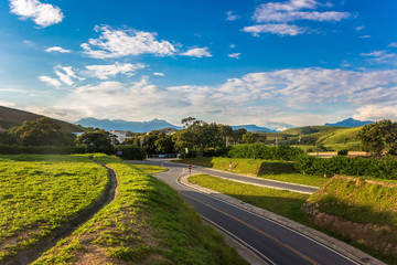 Fototapeta na wymiar Rural Road Among Grean Fields. Amazing Blue Sky With Beautiful Clouds, Mountains, Meadow With Fresh Green Grass. Highway In Rio de Janeiro District. Brazilian Countryside.