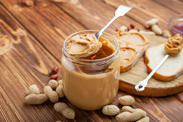 Peanut butter in spoon near creamy peanut paste in open glass jar, slice of peanut butter bread, toast. Peanuts in the peel scattered on the brown wooden table with copy space for cooking breakfast