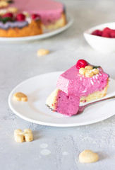 Piece of raspberry cream mousse cake (no baked cheesecake) decorated with fresh raspberries, mini cookies and coconut flakes on light grey background.