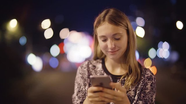 Close up of attractive woman using a smartphone. She is texting, taking a selfie, checking mails, chats or the news online. Night. City background.   