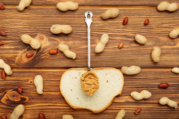 Vintage spoon with creamy peanut butter on a toast, slice of bread. Peanuts in the shell and peeled peanuts on brown wooden table. Flat lay of peanut paste for cooking breakfast. Vegan food concept.