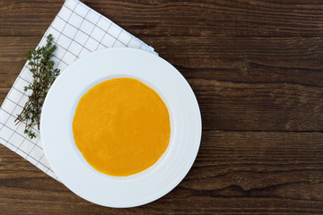 Pumpkin puree soup with a white ceramic plate on a wooden table. Next to a checkered napkin and a bunch of thyme. Copy space