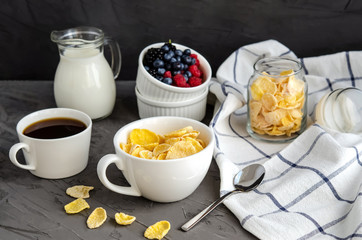 Healthy breakfast with cornflakes in a white cup, berries, milk and coffee on a dark gray background.