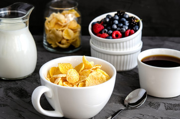 Healthy breakfast with cornflakes in a white cup, berries, milk and coffee on a dark gray background.