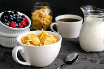 Healthy breakfast with cornflakes in a white cup, berries, milk and coffee on a dark gray background.