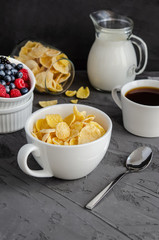 Healthy breakfast with cornflakes in a white cup, berries, milk and coffee on a dark gray background.