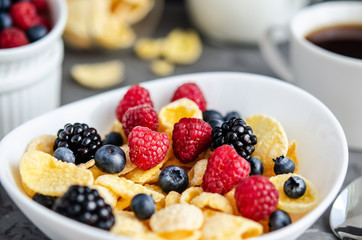 Healthy breakfast with cornflakes in a white plate, berries, milk and nuts on a dark gray background.