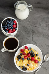 Healthy breakfast with cornflakes in a white plate, berries, milk and coffee on a dark gray background.
