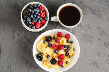 Healthy breakfast with cornflakes in a white plate, berries, and coffee on a dark gray background.