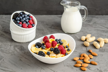 Healthy breakfast with cornflakes in a white plate, berries, milk and nuts on a dark gray background.