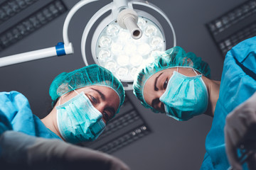 From below female surgeons in medical uniform using professional tools while standing under bright light in operating theater
