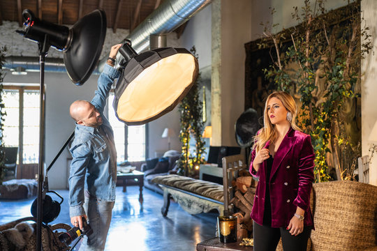 man setting lights of alluring adult lady in stylish colorful clothes sitting in armchair against vintage rustic interior among bright light of soft box and reflector in photo studio