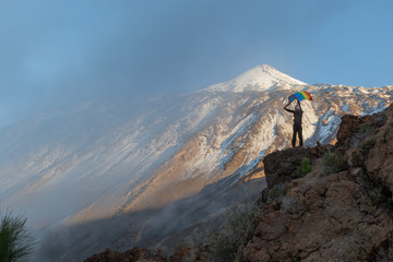 Active touristic woman in warm clothes climbing on top of mountain and waving colorful LGBT flag in stony peak in Tenerife, Canary Island, Spain