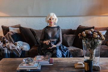 Gorgeous senior woman in elegant black dress with leather handbag looking at camera while sitting on brown velvet sofa with pet among cushions beside rustic wooden table with books and dry bouquet in vase in spacious living room of country house