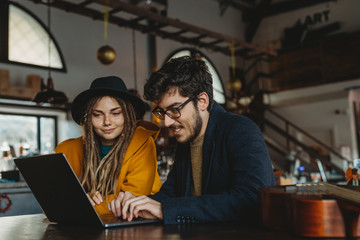 Smart man in glasses and stylish woman in hat looking at monitor while man typing on laptop in cafe