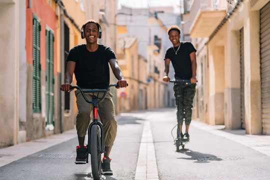 Cheerful young African American man riding electric scooter while black male is driving bicycle in street looking at camera