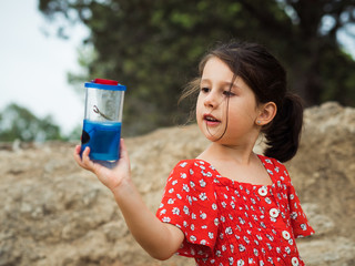 Little girl in red dress examining insect in jar on summer day in nature