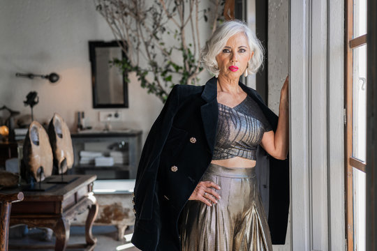 Low angle of confident elderly lady in stylish black jacket and golden shirt and trousers looking at camera while standing with hand on hip and leaning on window frame against blurred luxurious retro interior with antique furniture at home