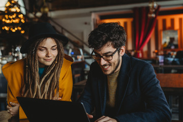Smart man in glasses and stylish woman in hat looking at monitor while man typing on laptop in cafe
