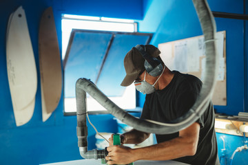 Worker in protective mask adjusting details surfboard in workshop