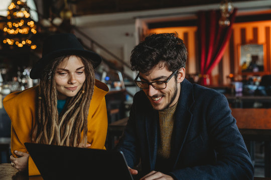 Smart man in glasses and stylish woman in hat looking at monitor while man typing on laptop in cafe