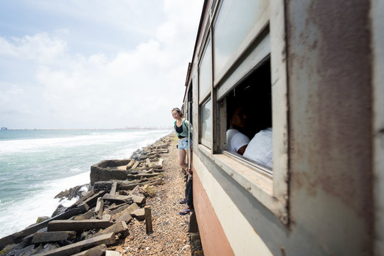Side view of resting female in casual wear looking at camera from doorway of train going along seashore at Sri Lanka 