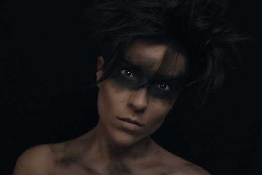 Pensive young dark haired female model with uncombed hair and black make up around eyes looking at camera in studio on black background