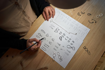 Cropped unrecognizable woman jewelry designers with a pencil in hand making jewelry drawings on a paper on a wooden table