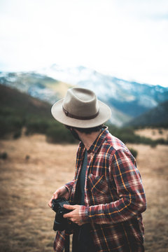 Stylish traveler in hat and shirt with photo camera looking back on stony mountains with tree