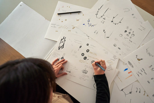 From above cropped unrecognizable woman hands sitting at a table with a pencil drawing jewels on paper
