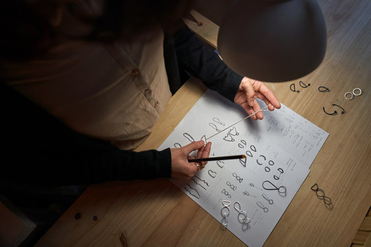 Cropped unrecognizable woman jewelry designers with a pencil in hand making jewelry drawings on a paper on a wooden table