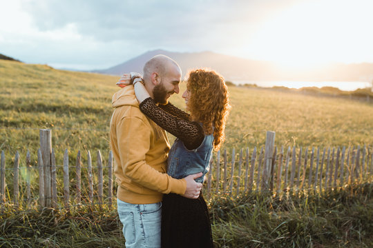 Young bald bearded man in yellow hoodie and jeans shorts embracing happy curly haired girlfriend in dress and denim vest while standing near fence together on meadow