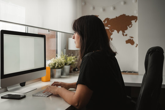 Side view of concentrated dark haired female employee focusing on screen and typing on computer while sitting at table in light office in Paris 