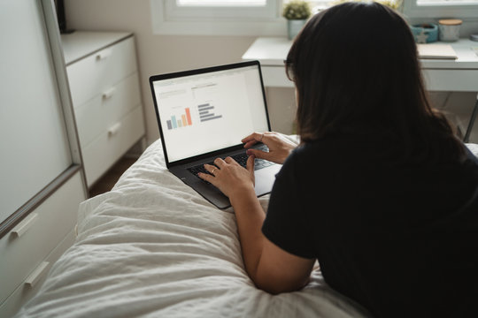 from above view of concentrated young woman in glasses typing on laptop while studying and laying on bed at home in Paris