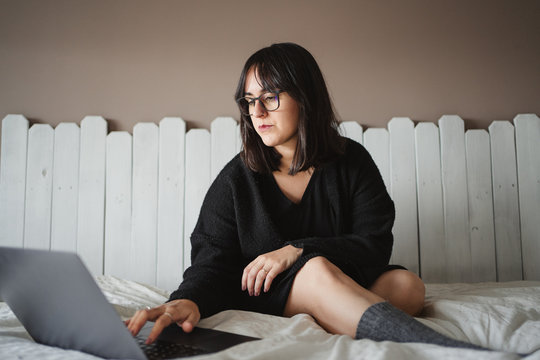 view of concentrated young woman in glasses typing on laptop while studying and laying on bed at home in Paris