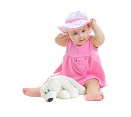 A little baby girl in pink dress and panama hat sits on white background in studio with little bear cub, isolated