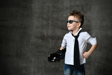 Smiling boy in stylish casual white shirt, jeans, tie and sunglasses standing and holding black toy car present in hand