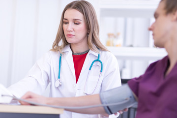 A female doctor wearing a white coat with a stethoscope is checking the blood pressure of the patient.