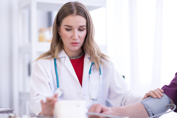 A female doctor wearing a white coat with a stethoscope is checking the blood pressure of the patient.