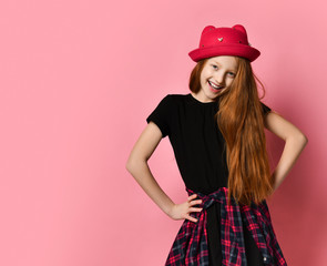 Teen female in black dress, checkered shirt on waist, red hat. She is laughing, hands on hips, posing on pink background. Close up