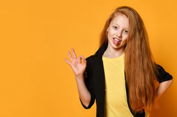 Red-headed teenager in black jacket and yellow t-shirt. She is showing tongue and okay sign, posing on orange background. Close up