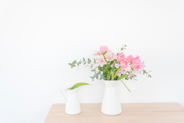 Fototapeta na wymiar 白い花瓶とピンクの花　テーブル　木製テーブル　白い壁　室内　部屋　屋内　アレンジメント　花束　花　春　ピンク　観葉植物　華やか　爽やか　春らしい　かわいい　おしゃれ　白背景　文字スペース　コピースペース　ホワイトスペース　余白　横位置