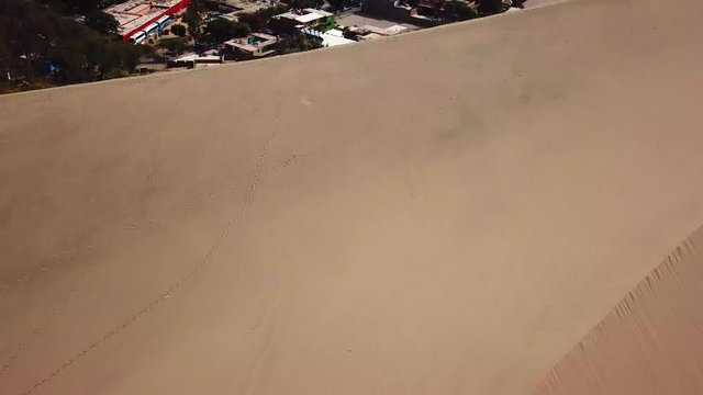 Aerial, til up, drone shot, over sand dunes, revealing the Huacachina lagoon and the oasis village, at Atacama desert, on a sunny day, in south Peru